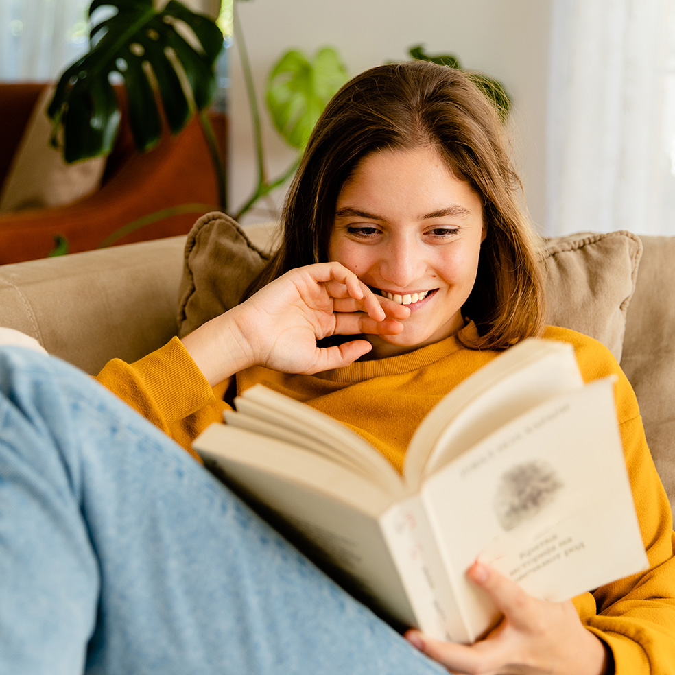Ein Mädchen im Teenageralter sitzt zuhause auf dem Sofa und liest ein Buch.