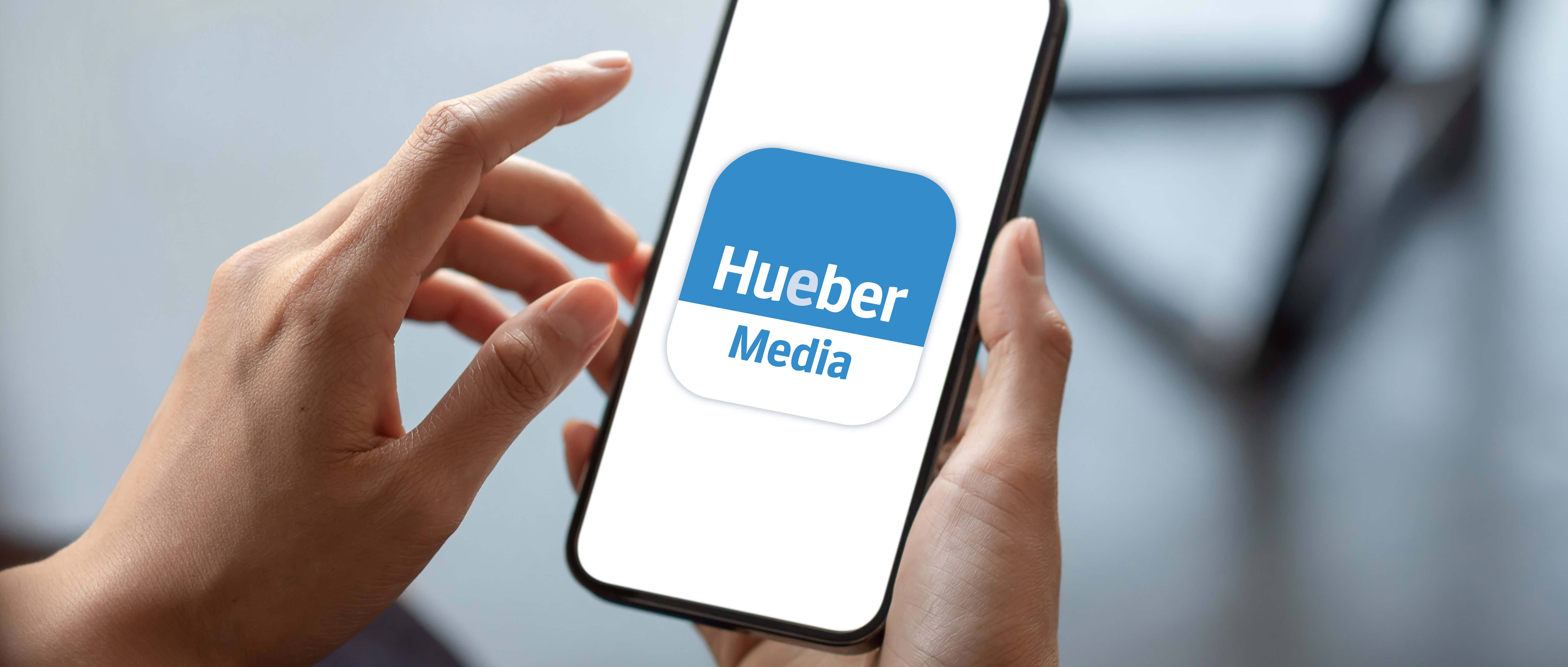 Zwei Hände halten ein Smartphone auf dem das blau-weiße Logo der Hueber Media App zu sehen ist.